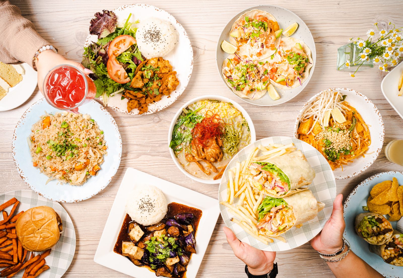 A table spread of plant-based dishes: fried rice, ramen, pad thai, tacos, wraps with fries, eggplant and tofu with rice, a burger with sweet potato fries and more